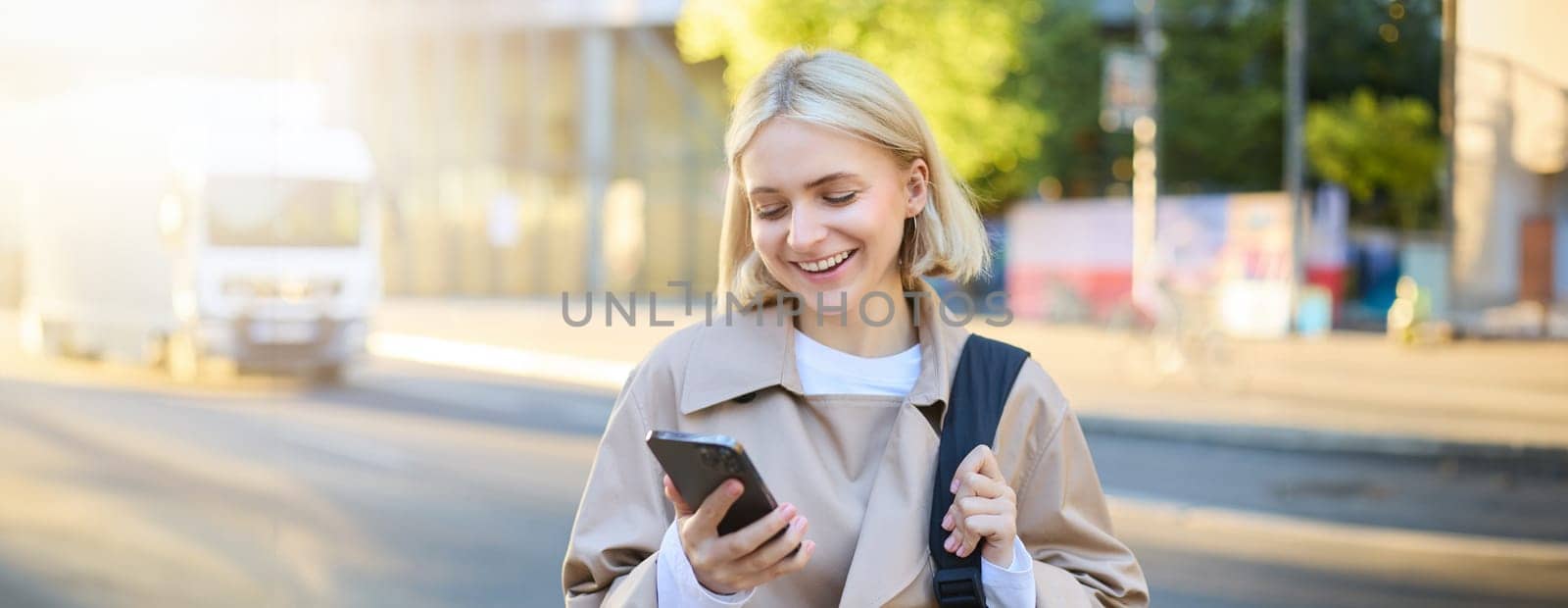 Close up portrait of happy, beautiful young woman with backpack, standing on street and using mobile phone, waiting for taxi near road, smiling by Benzoix