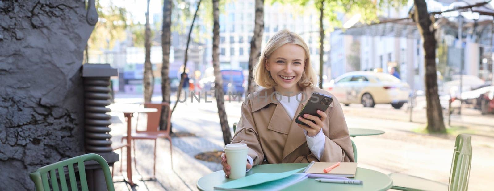 Portrait of blond smiling woman with smartphone, holding cup of coffee, drinking chai and enjoying sunny day outdoors in city centre.