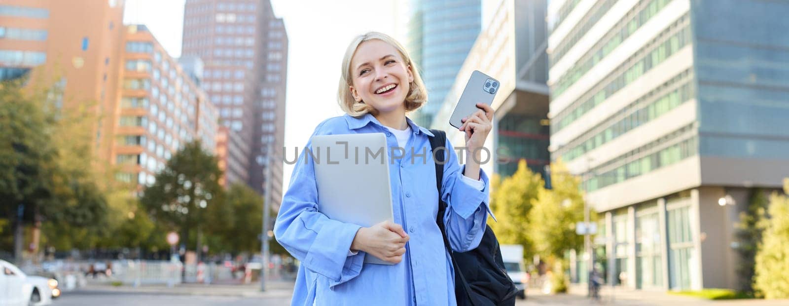 Image of young stylish, modern blond woman in blue shirt, holding laptop and backpack, using smartphone, smiling, walking along the street, posing in city centre by Benzoix