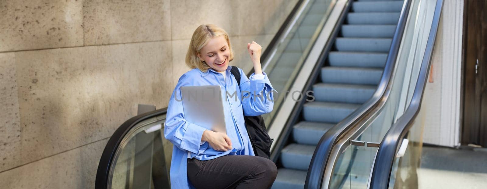 Excited young blond woman with backpack and laptop, says yes, winning, celebrating victory, achieve goal, posing near escalator by Benzoix