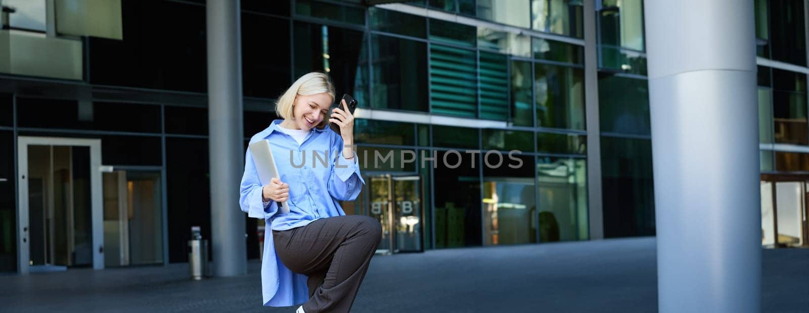 Vertical shot of stylish, happy modern blond girl, student dancing with smartphone near office building, saying yes, making fist pump, celebrating, feeling excitement.