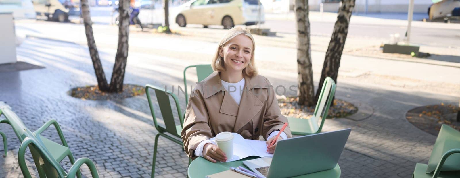 Portrait of young woman working on project, sitting outdoors in cafe, drinking her coffee, using laptop and making notes in notebook, studying, preparing for work interview.