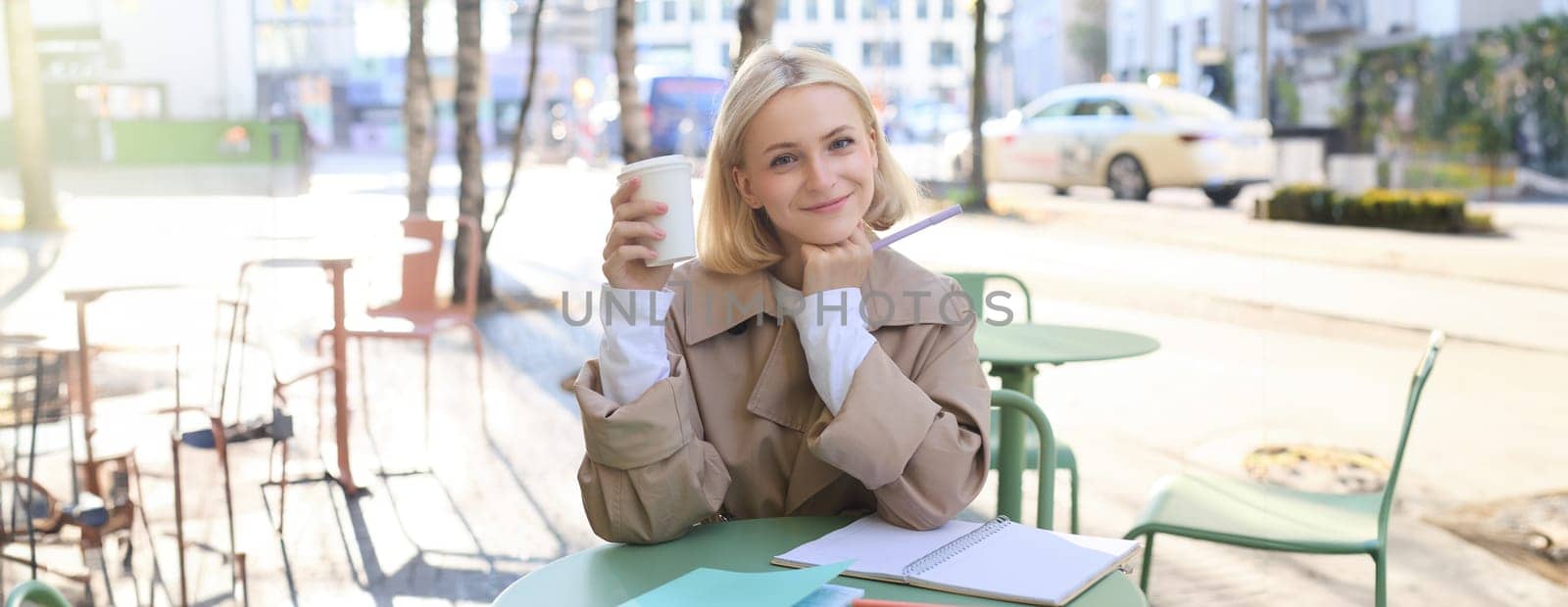Stylish young urban woman, sitting in cafe and drinking coffee, smiling and looking at camera, doing homework or working on project, making notes in notebook.