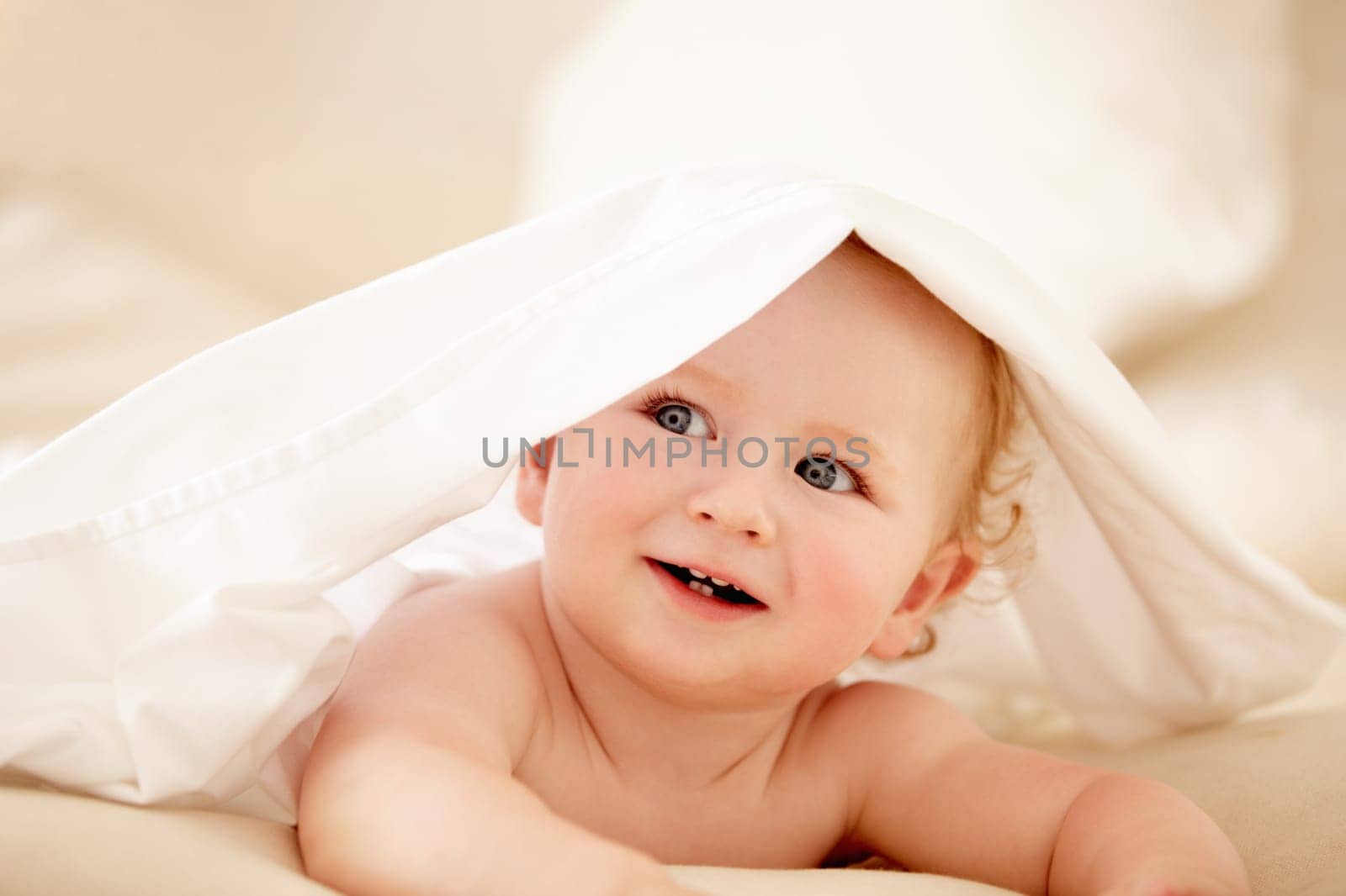 Smile, face and happy baby on a bed with blanket for playing, games or fun in a nursery room. Learning, child development and curious little boy kid in a bedroom with sheet cover while lying in house by YuriArcurs