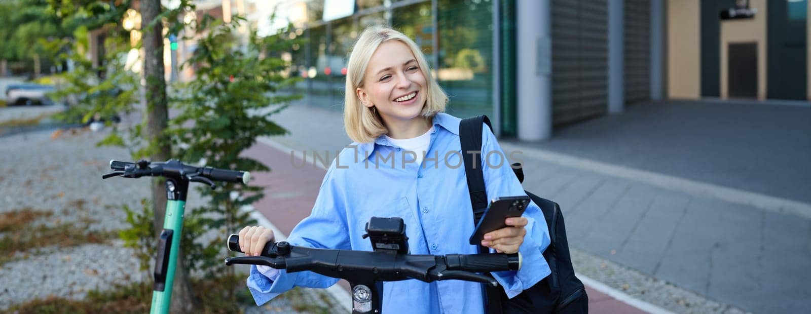 Portrait of smiling blond woman, college student using smartphone app to scan QR code on green electric scooter on street, renting a ride.