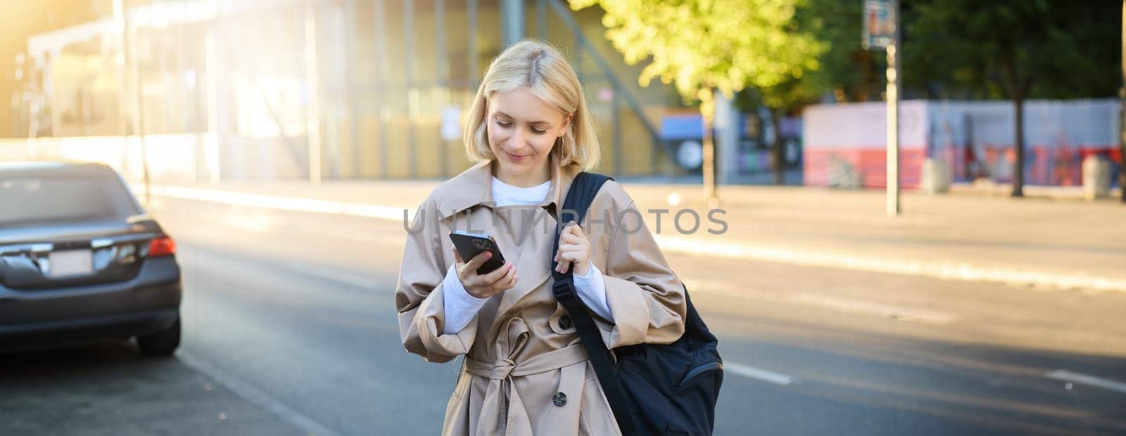 Image of young modern woman, student waiting for someone on street, standing with backpack and checking her mobile phone, looking at smartphone screen.