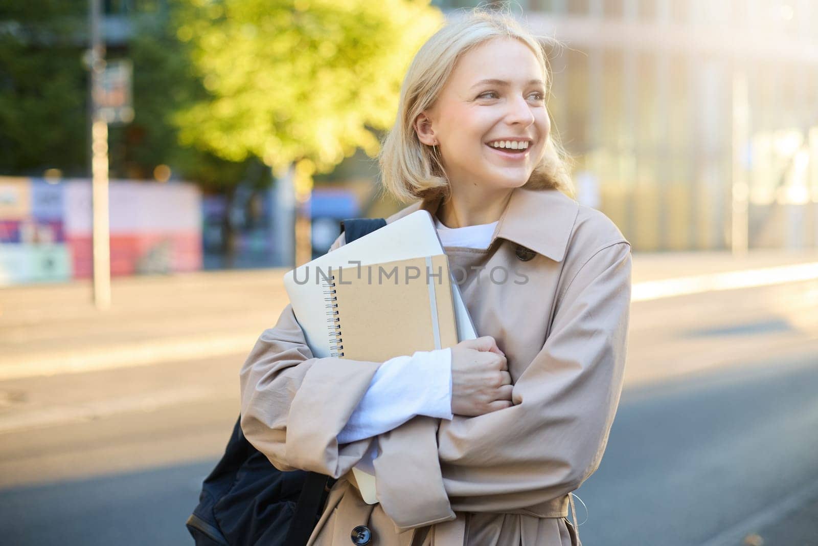 Candid young woman on her way to University, standing on street, carries notebooks and homework, has backpack on shoulder, turns away, laughs and smiles, shows genuine emotions by Benzoix
