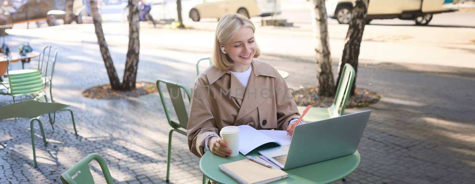 Portrait of young blond woman, female student in street cafe, wearing wireless headphones, using laptop, having online meeting, attend web lecture or course, doing homework by Benzoix