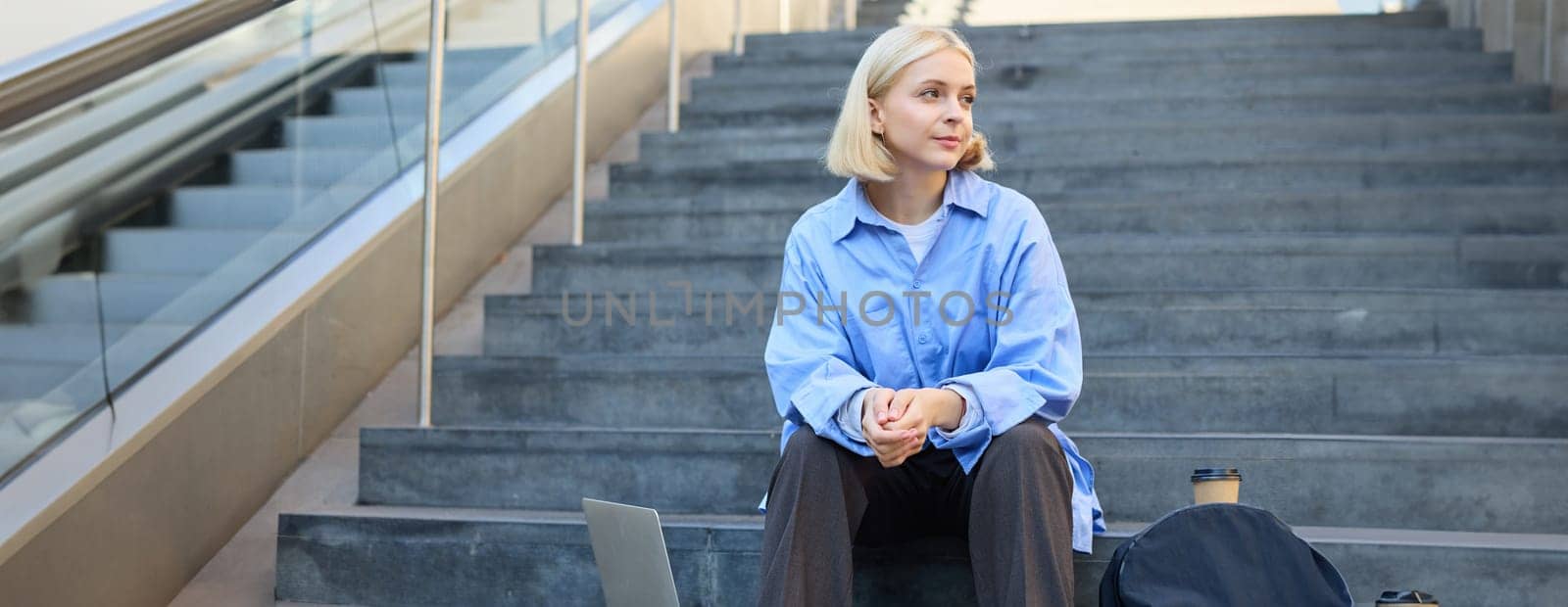 Urban style portrait of young woman, student sitting on city street stairs, with backpack, cup of coffee and laptop, looking happy and upbeat, smiling.