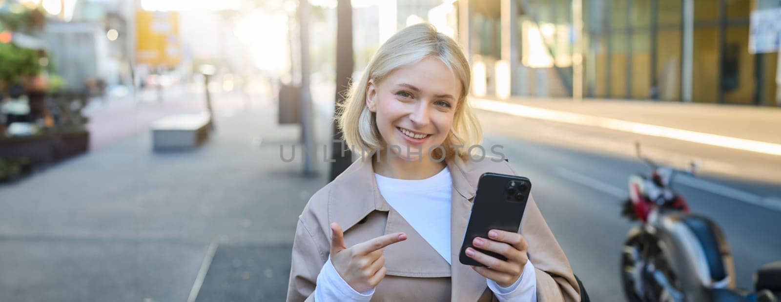 Close up portrait of young blond woman, student pointing at smartphone, standing on street and showing smth on mobile phone by Benzoix