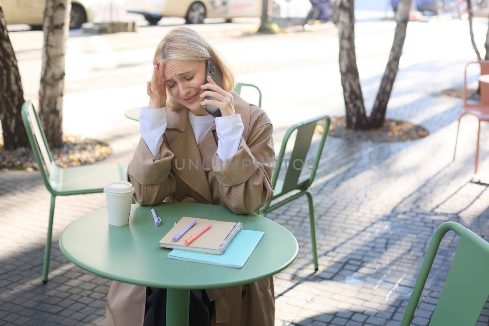 Image of young stressed, upset woman, talking on mobile phone with worried face expression, has difficult conversation over the telephone, sitting in outdoor cafe by Benzoix