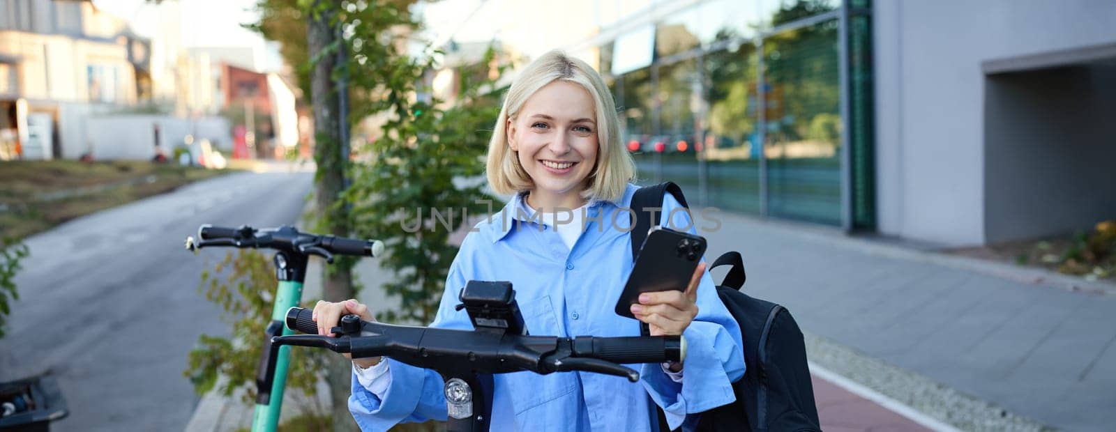 Portrait of young smiling woman, unlocks electric scooter, scans qr code with smartphone app to unlock her ride, standing on street near building.