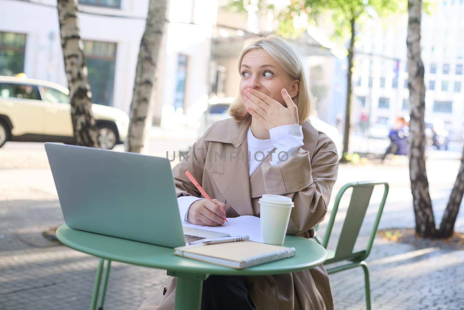Image of woman sitting with laptop outside on street, using laptop, looking surprised, amazed by something she saw online by Benzoix