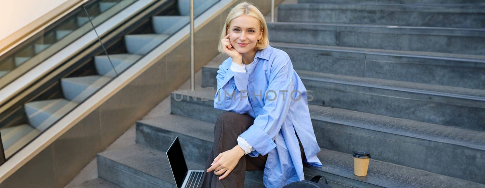 Portrait of young urban woman, college student with backpack, laptop and coffee, sitting on stairs in city, resting outdoors, taking break.