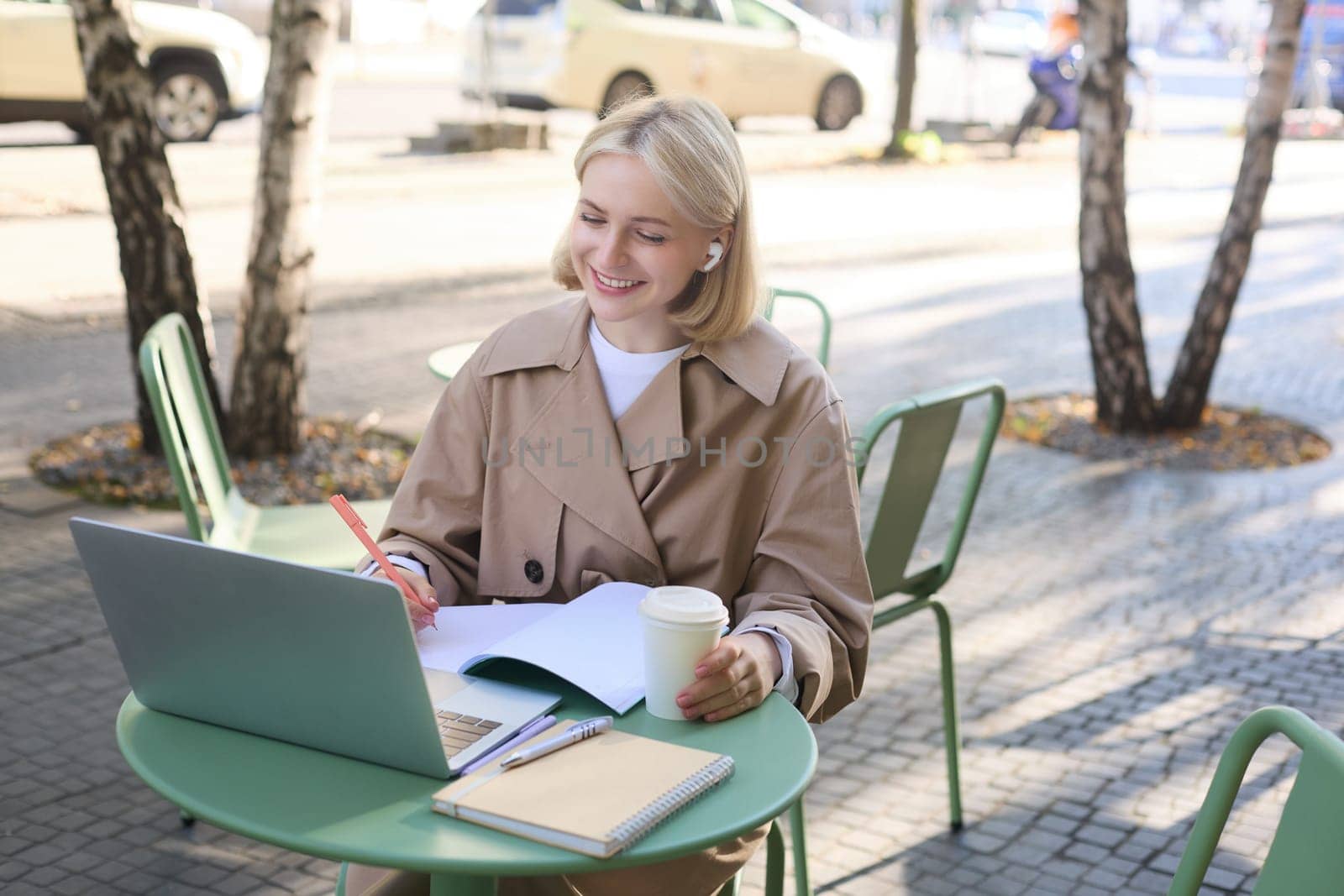 Portrait of young blond woman, female student in street cafe, wearing wireless headphones, using laptop, having online meeting, attend web lecture or course, doing homework by Benzoix
