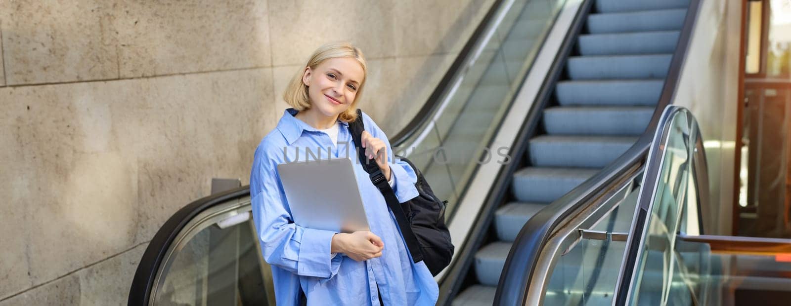Image of happy, positive young woman in blue shirt, holding laptop and backpack, posing near escalator, tilt her head and smiling at camera.