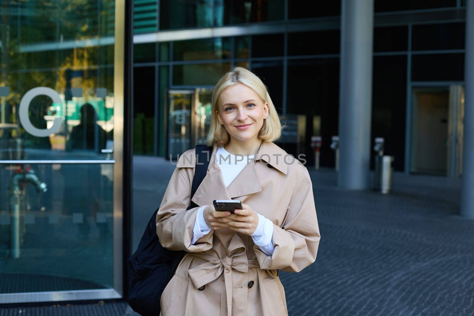 Street photo of young blond woman with smartphone, walking in city, waiting for someone, order taxi on mobile application by Benzoix