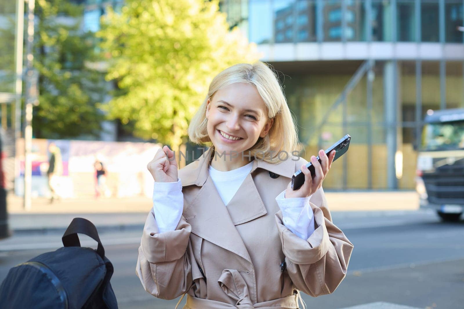 Cheerful blonde woman, sitting on bench and celebrating victory, holding mobile phone, smiling pleased, triumphing with joy, receive good news.