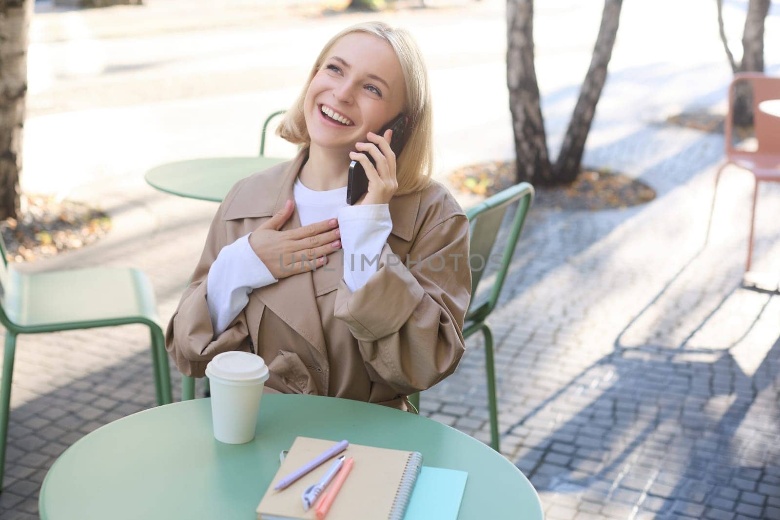 Lifestyle and people concept. Happy beautiful young woman, talking on mobile phone, sitting outside in cafe with cup of coffee, smiling and laughing, chatting about something funny by Benzoix
