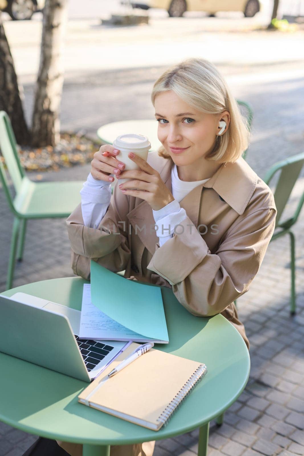 Vertical shot of cute blond woman, sitting in street cafe outdoors, wearing wireless headphones, using laptop, drinking her coffee and smiling at camera.