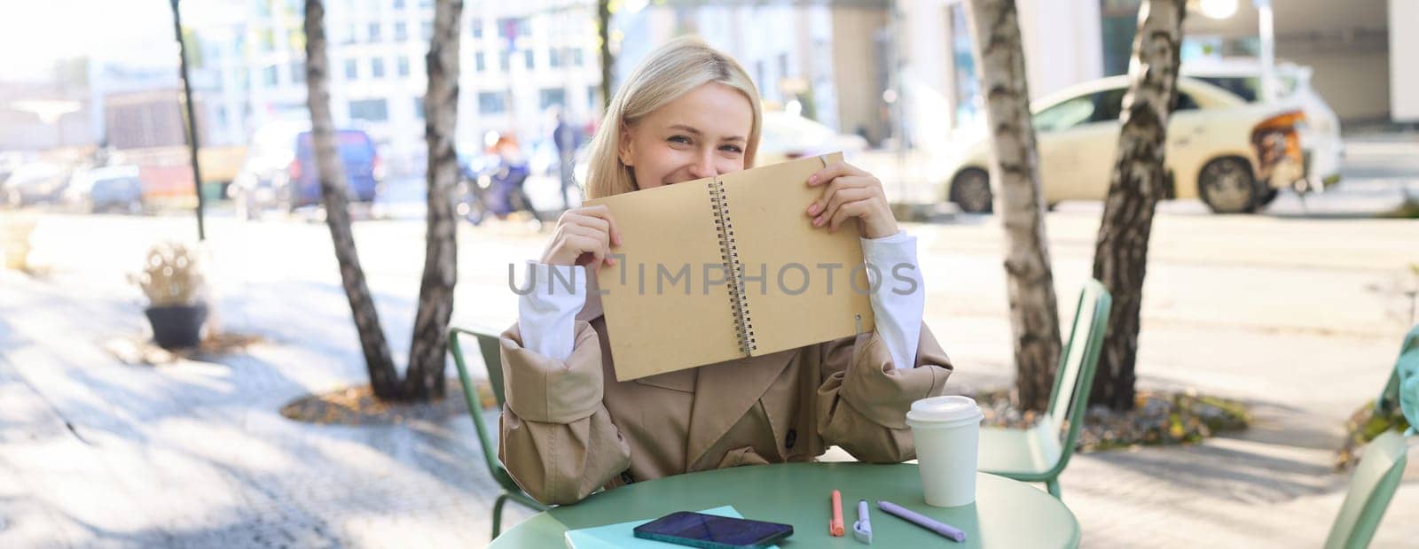 Image of young smiling blond woman sitting in an outdoor cafe, holding notebook, doing her homework outside in coffee shop, looking happy.