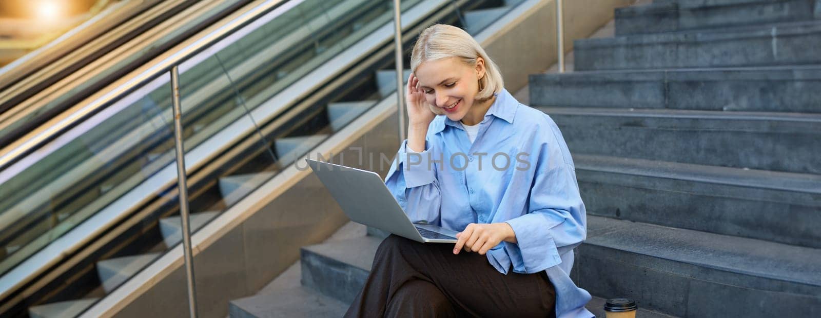 Image of young woman, student studying online, working remotely, sitting on stairs with laptop and drinking coffee in takeaway cup, elearning.