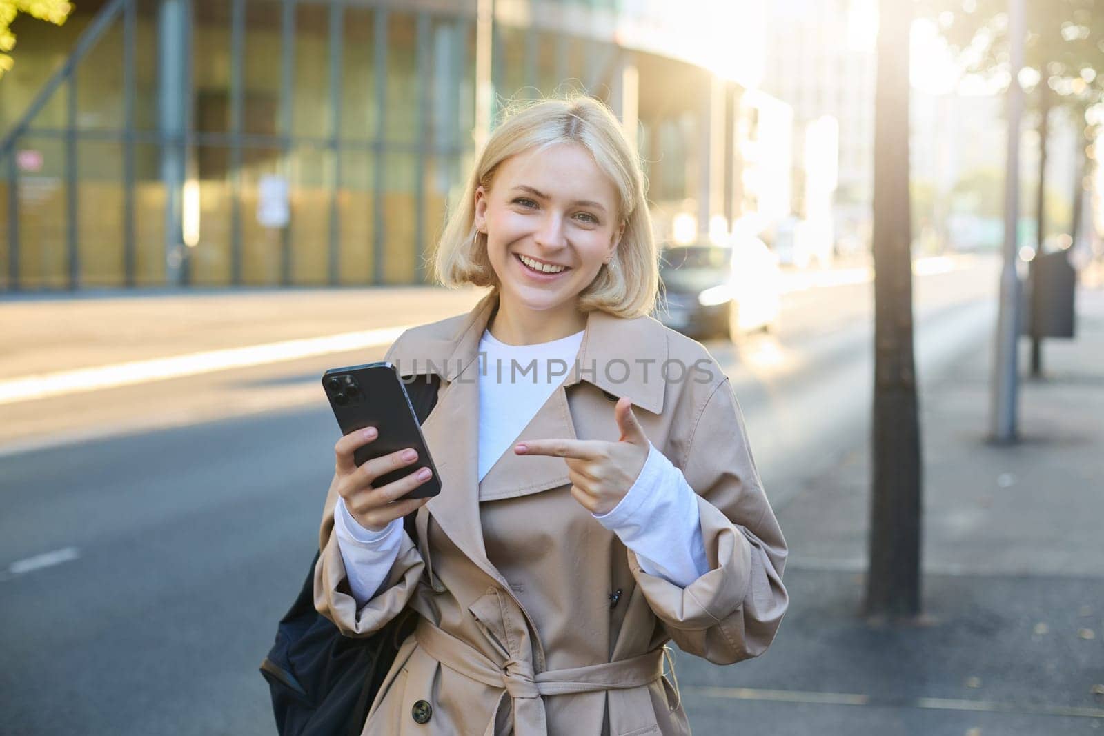 Portrait of young woman on streets of city, holding smartphone, pointing at her mobile phone, looking happy and smiling at camera.