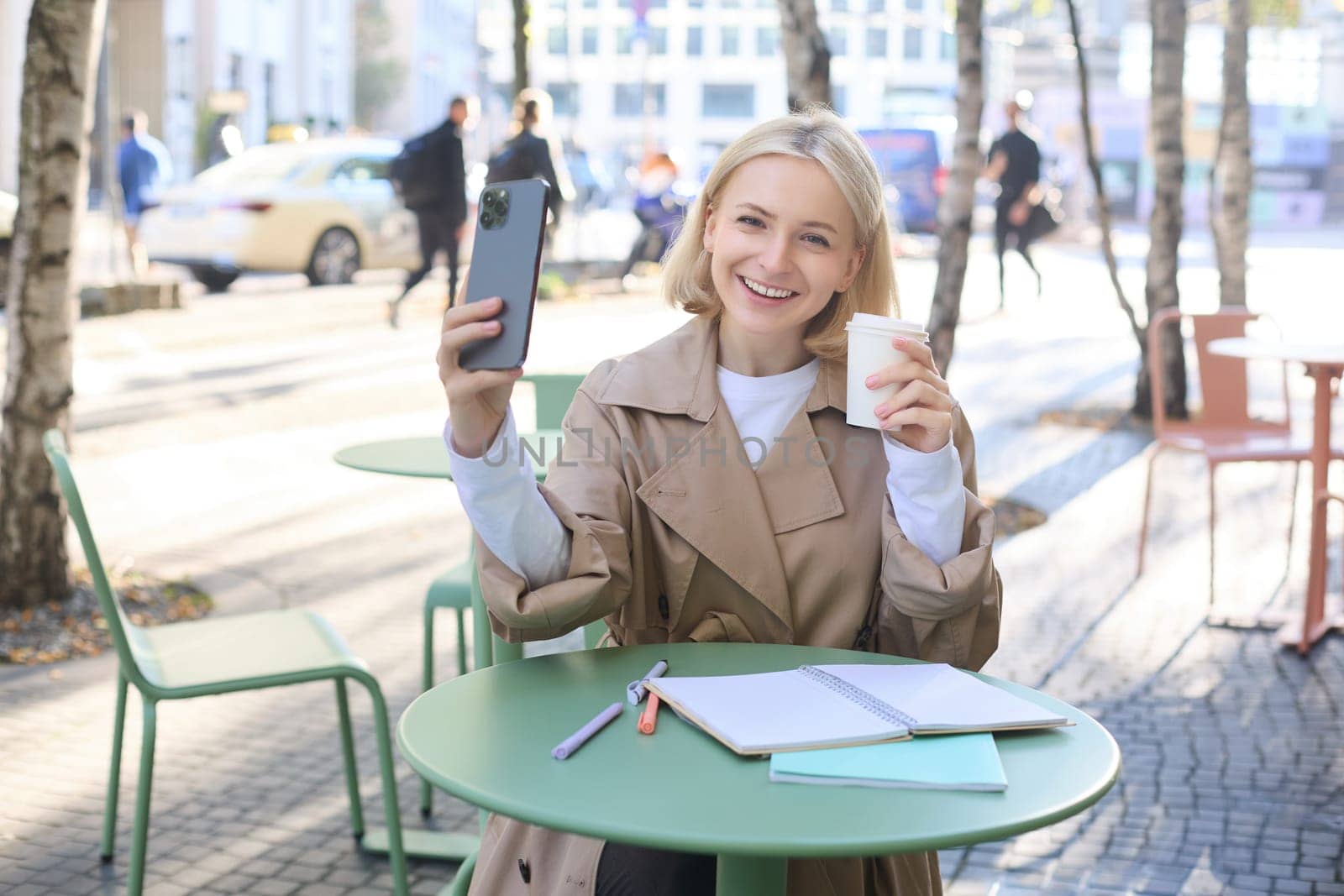 Lifestyle shot of stylish blond girl with smartphone, drinking coffee, posing with her chai for social media, creating content on phone, taking selfie by Benzoix