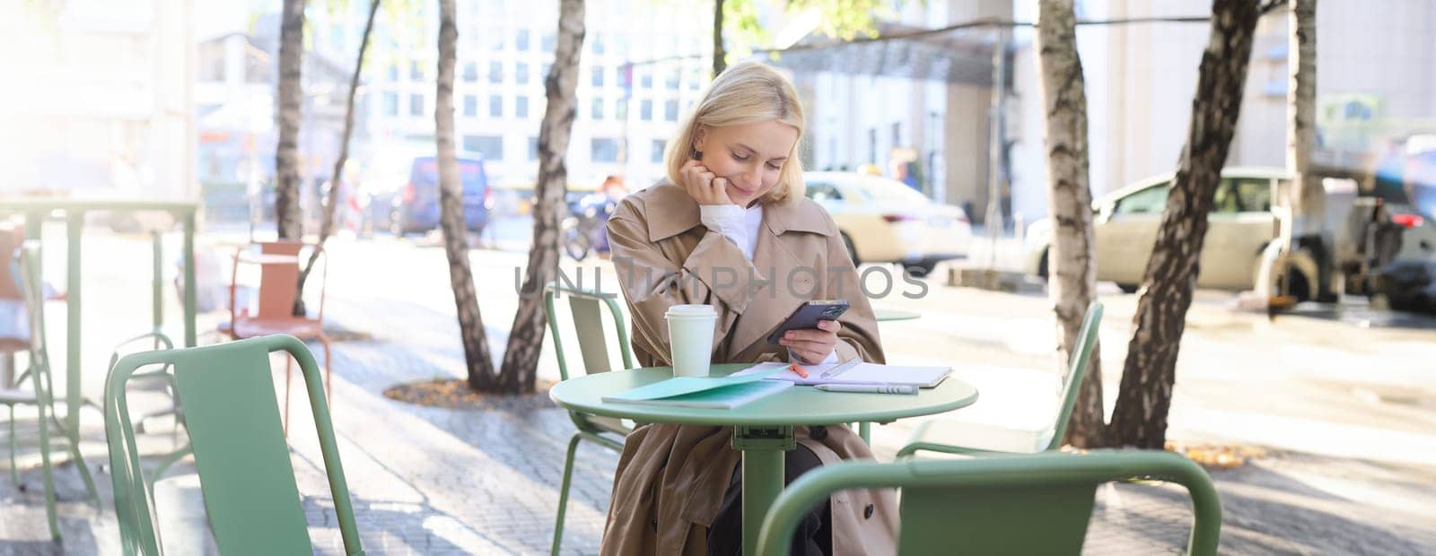 Portrait of modern young woman in city centre, sitting in cafe outdoors, drinking coffee, using mobile phone, smiling.