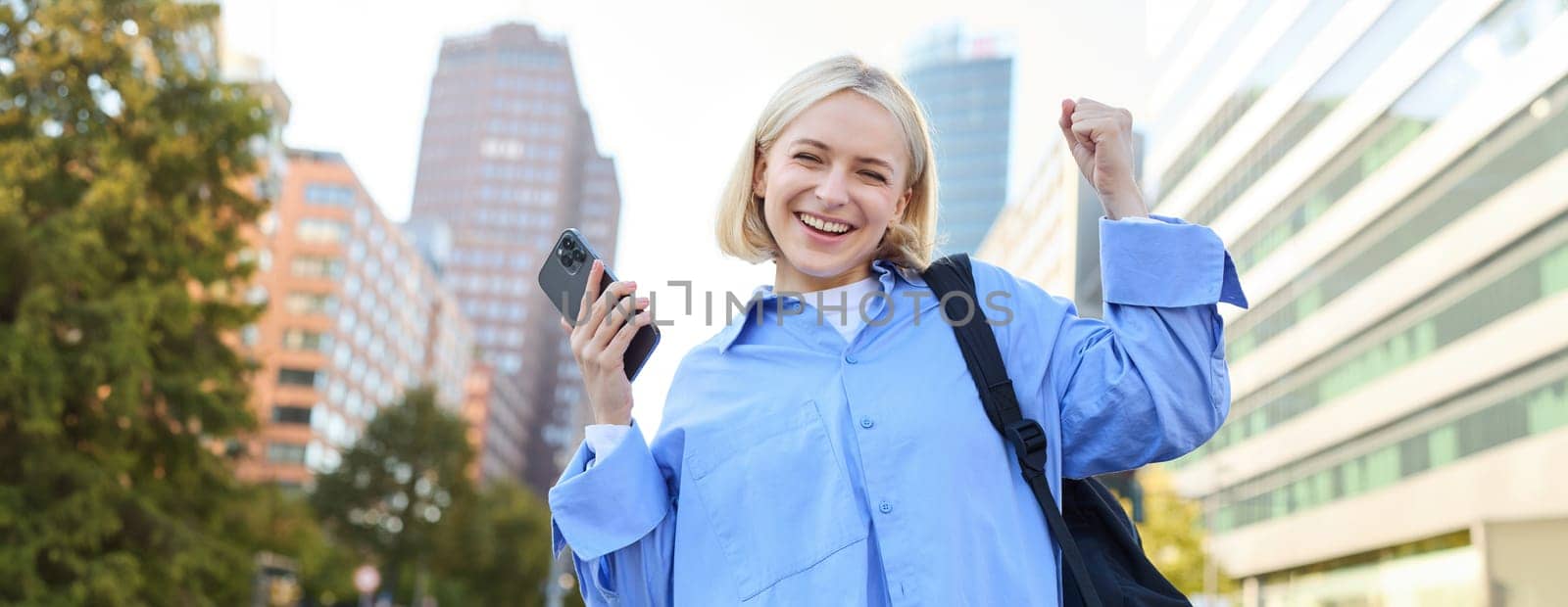 Confident, happy young woman celebrating, raising hand up and looking satisfied, using smartphone, triumphing while standing on empty street by Benzoix