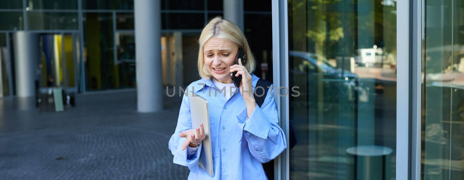 Happy female model, blond woman talking on mobile phone, chatting with someone, standing near office with upset, worried face, being in trouble.