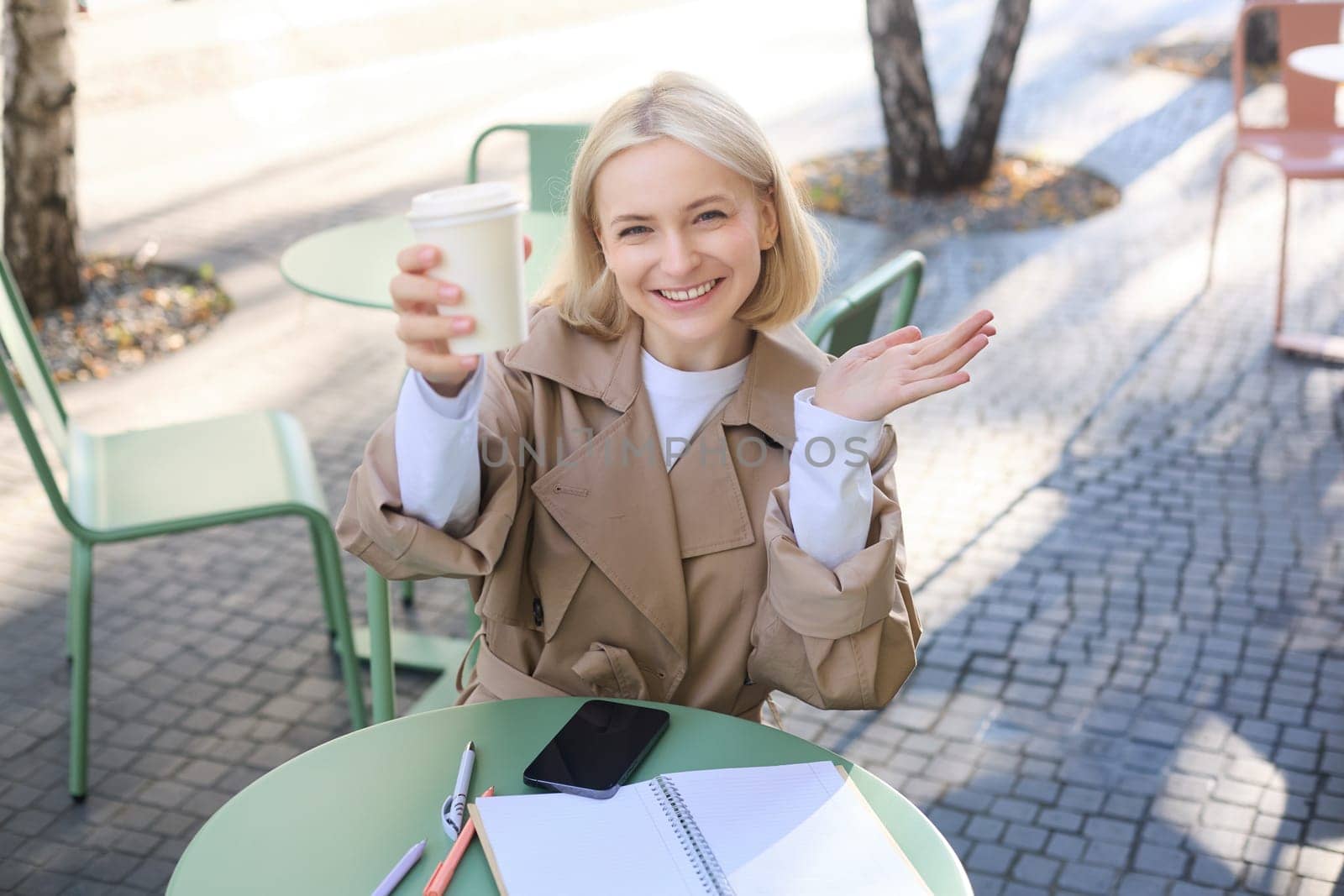 Food and drink concept. Young happy woman sitting in cafe, enjoying bright day outdoors, raising cup of coffee and smiling, drinking chai.