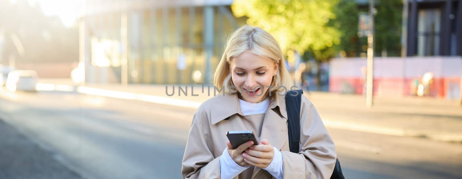 Portrait of surprised blonde woman, standing on street, looking at her mobile phone with amazed face, reading great news on smartphone by Benzoix