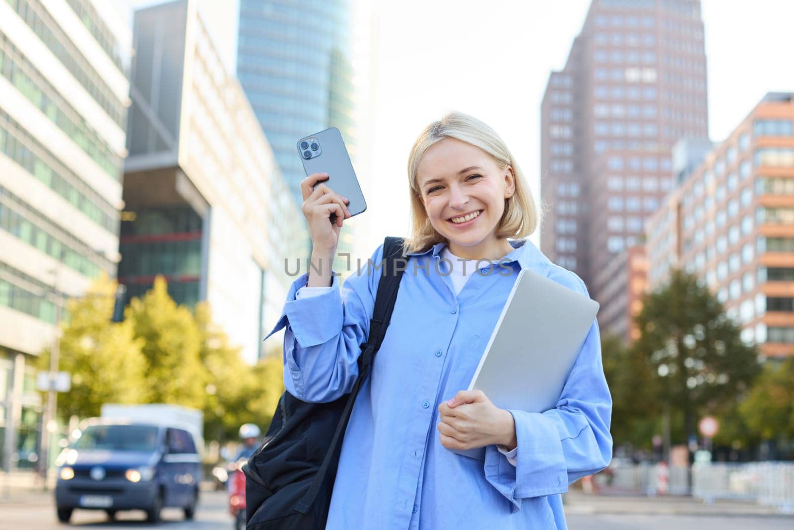 Image of young stylish, modern blond woman in blue shirt, holding laptop and backpack, using smartphone, smiling, walking along the street, posing in city centre by Benzoix