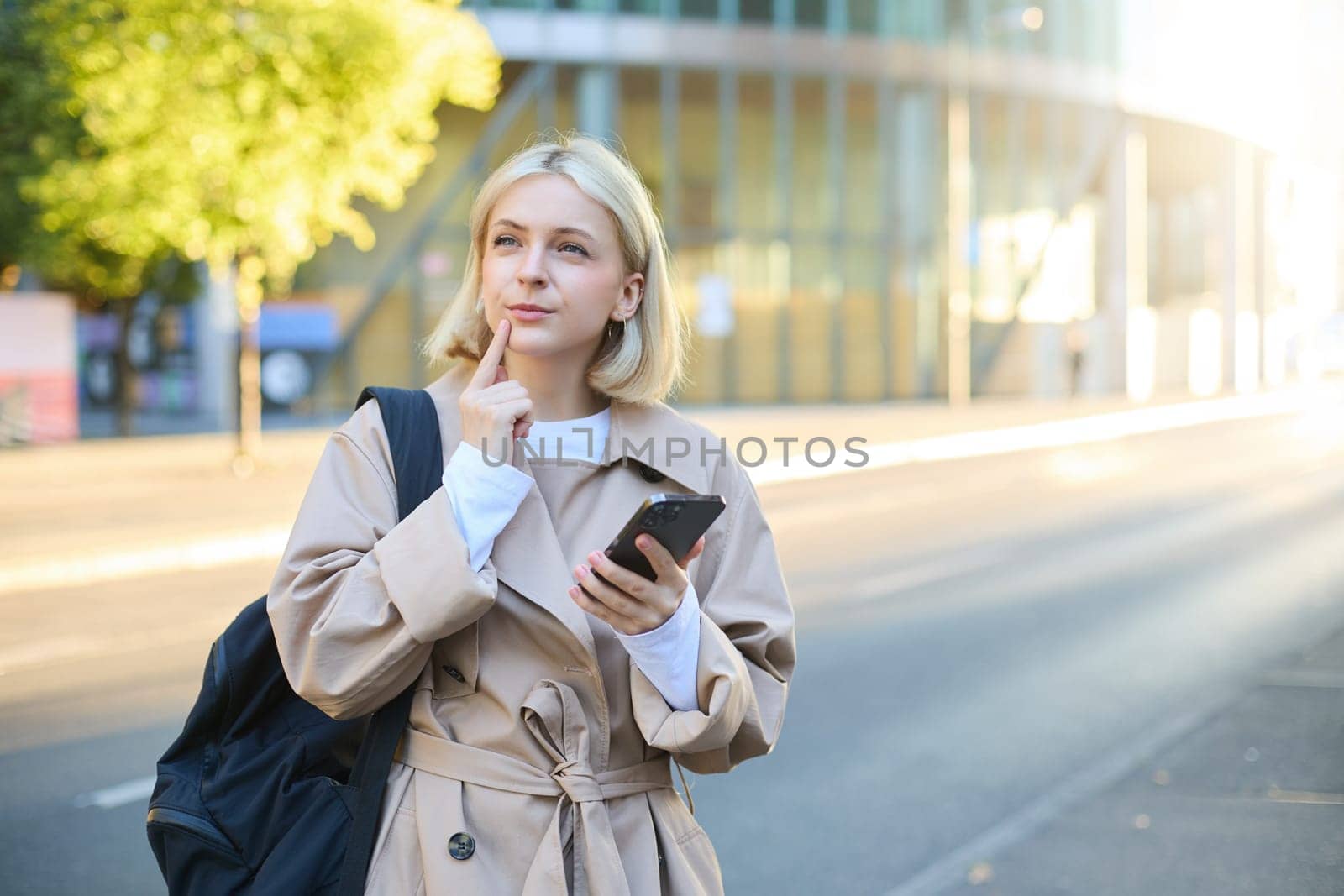 Portrait of woman with thinking face, standing with backpack on street, holding smartphone, pondering, making a decision with thoughtful face.