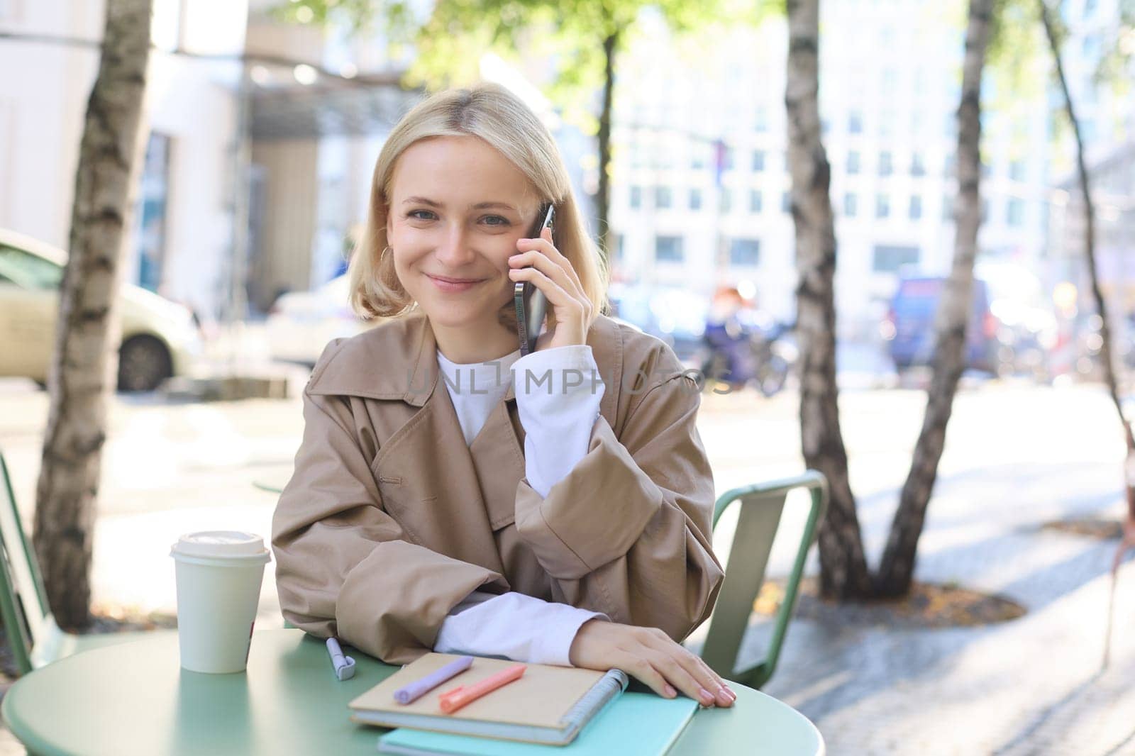 Outgoing young happy woman, sitting in cafe outdoors, talking on mobile phone, smiling, spending time outside in city centre, chatting. Lifestyle and people concept