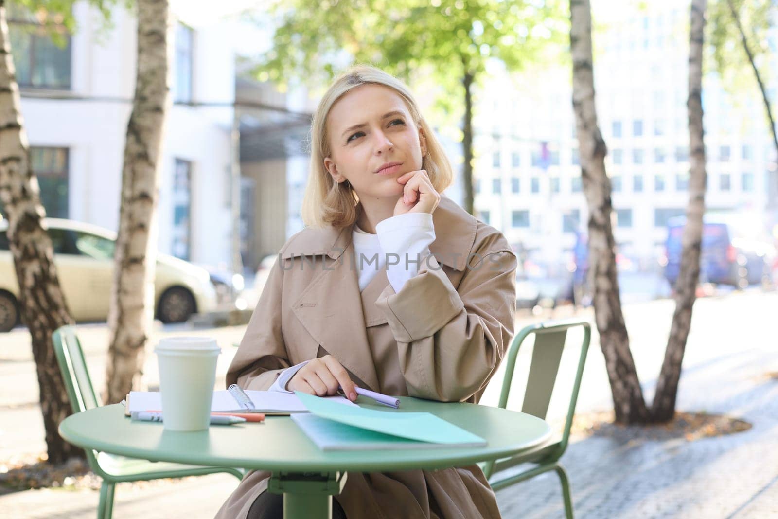 Portrait of thoughtful young woman, writing in her documents, making corrections, working on mistakes in homework, sitting in cafe with cup of coffee, drinking and studying.