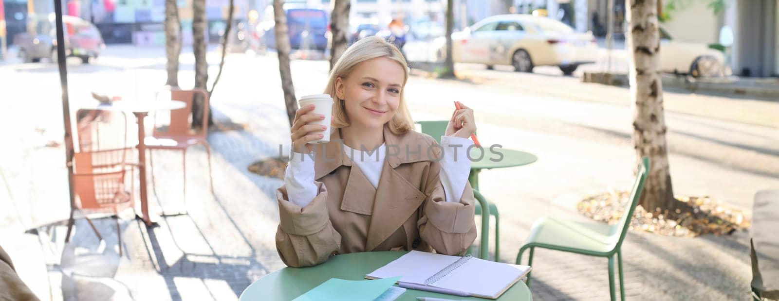 Image of beautiful young blond woman, girl drinking coffee in outdoor cafe, working on university project, doing homework, enjoying sunny weather, smiling at camera.