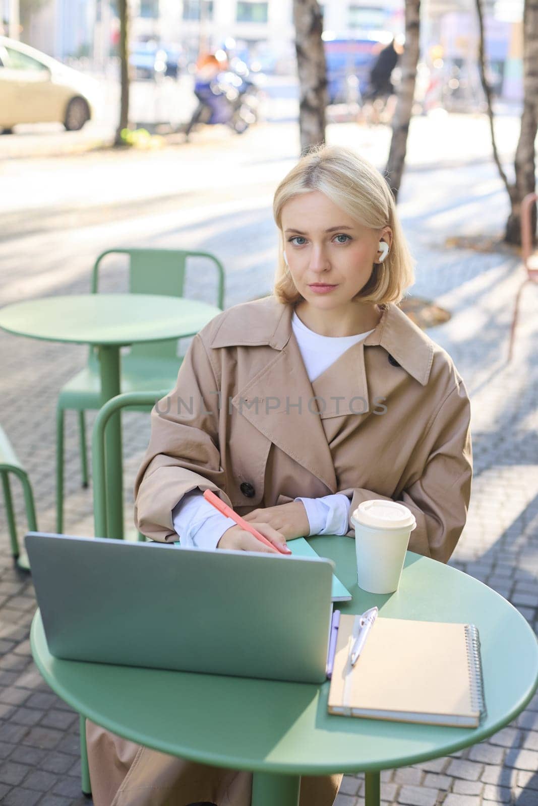 Vertical shot of young blond woman in wireless headphones, making notes, studying online, attend web lecture, using laptop, writing in notebook, looking serious at camera.