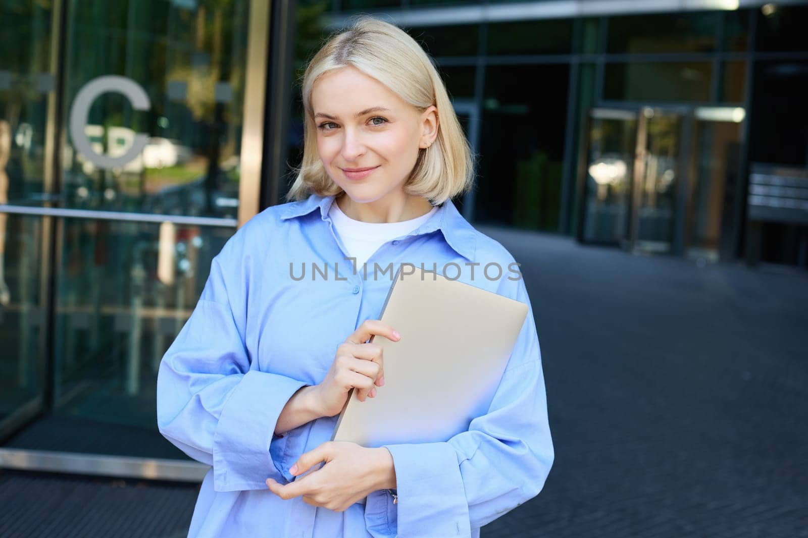 Image of young female employee, working woman standing near her office on street, holding pile of documents and smiling by Benzoix