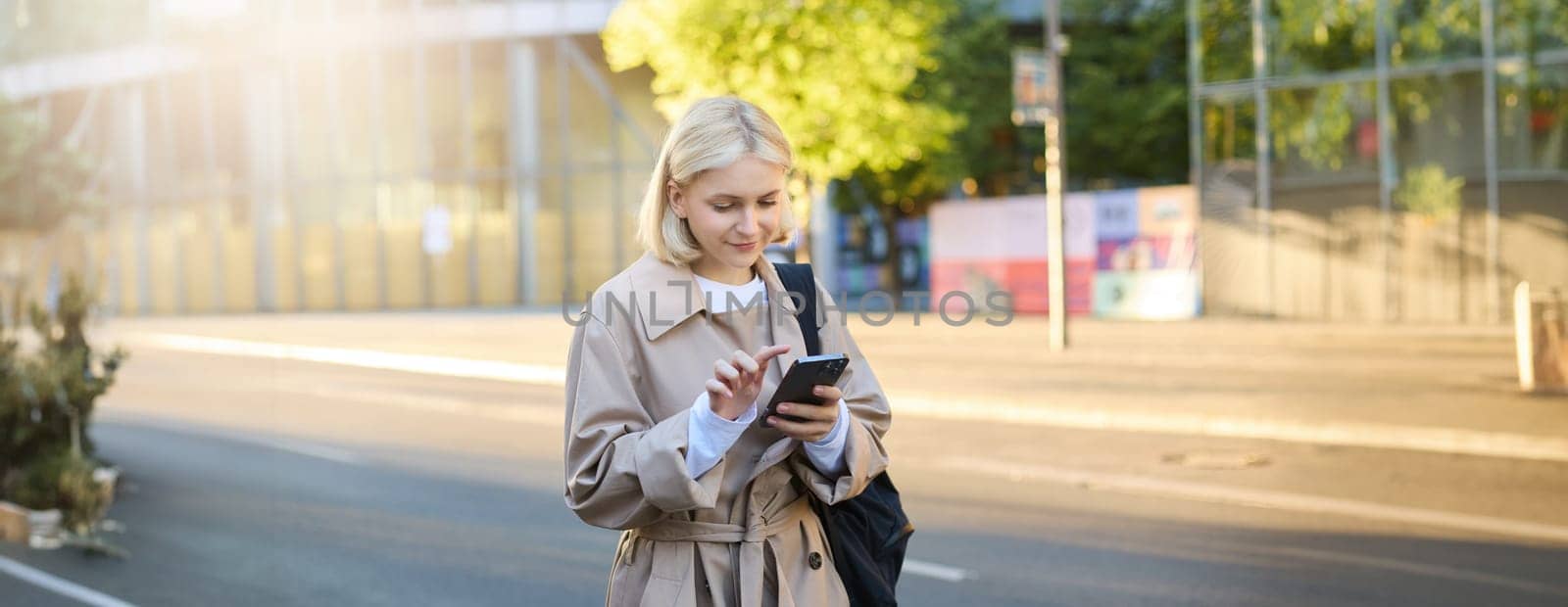 Image of young woman on street, carries backpack, looks at her smartphone, using carsharing application, orders taxi, sends a message on mobile app.