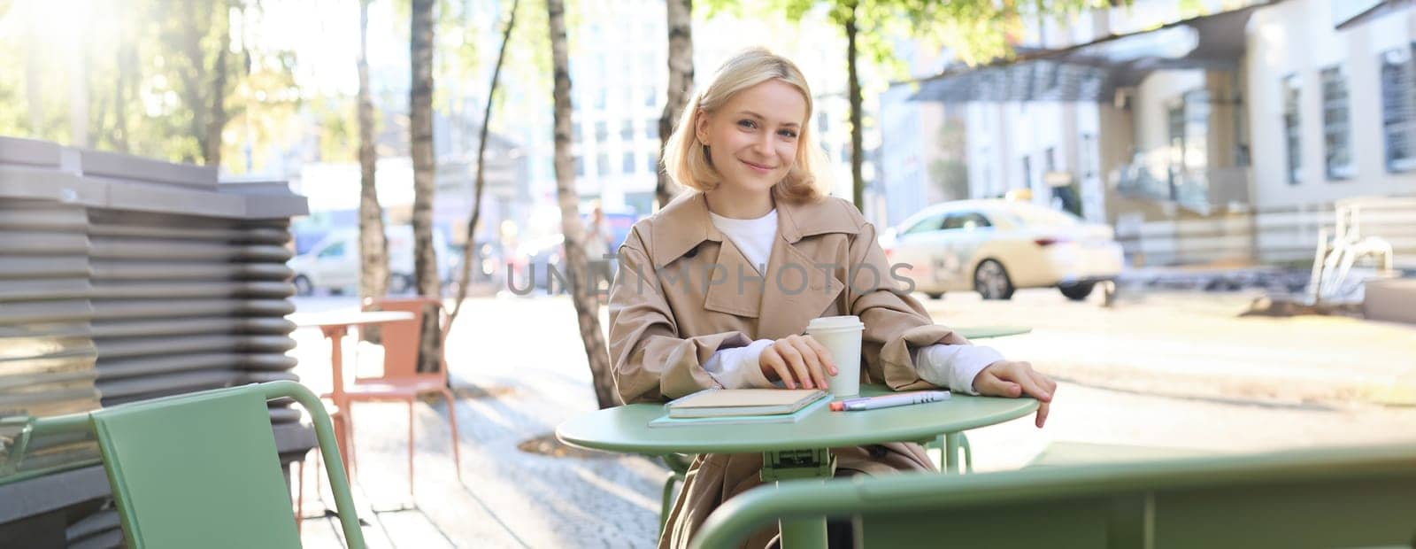 Portrait of young blonde woman sitting in cafe, doing homework and drinking coffee, writiing in journal, using notebook on fresh air.