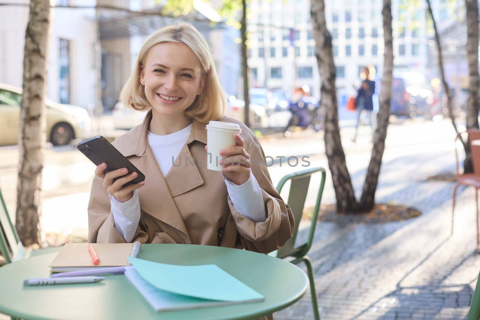 Portrait of blond smiling woman sitting in cafe, enjoying cup of takeaway coffee, using mobile phone, working on smartphone, social media blogger making post online by Benzoix