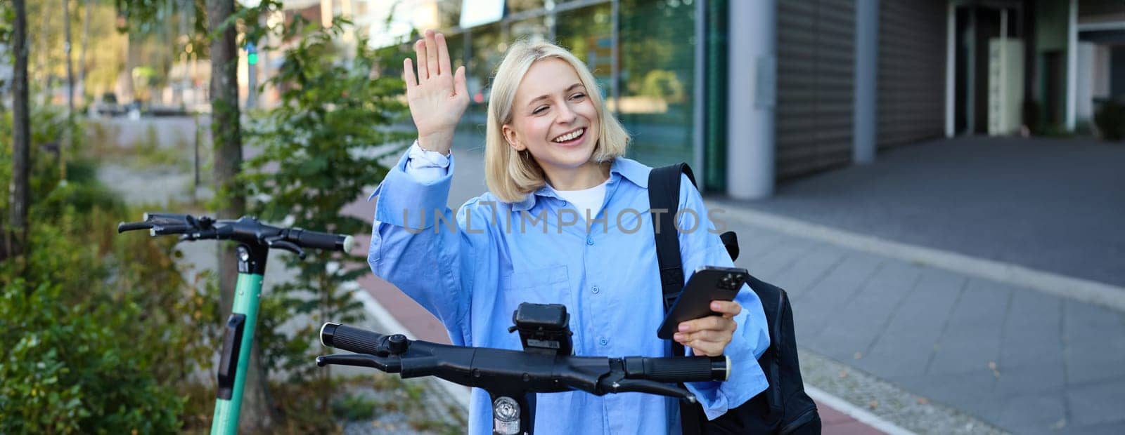 Young cheerful woman, tried to scan QR code on electric scooter on street, using mobile phone to rent it, saying hello to friend and waving hand at someone.