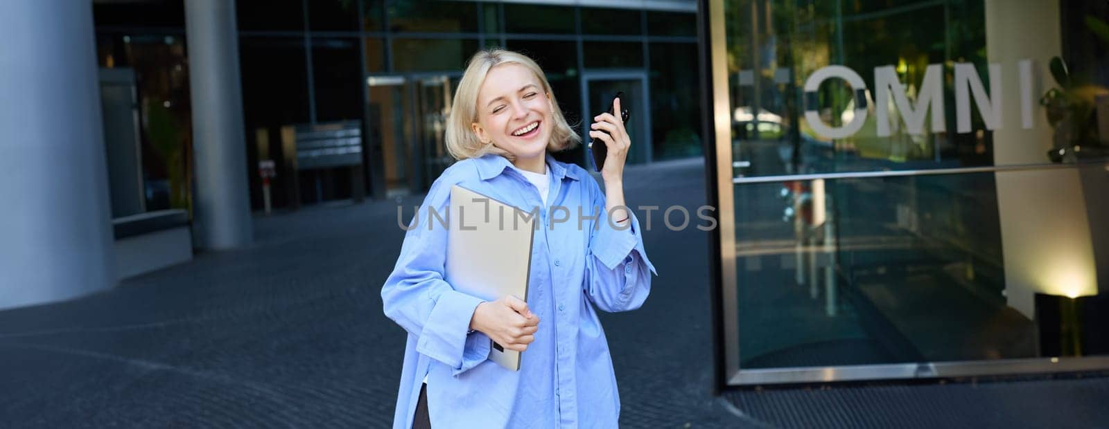 Carefree blond woman with laptop and smartphone, singing, listening music on mobile phone app, posing near office building or college campus on street.