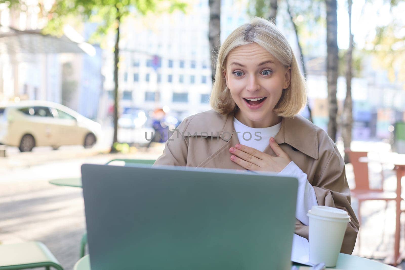 Lifestyle portrait of young surprised woman, looking at laptop with amazed face, sitting in coffee shop with excited face expression, spending time outdoors on sunny day by Benzoix