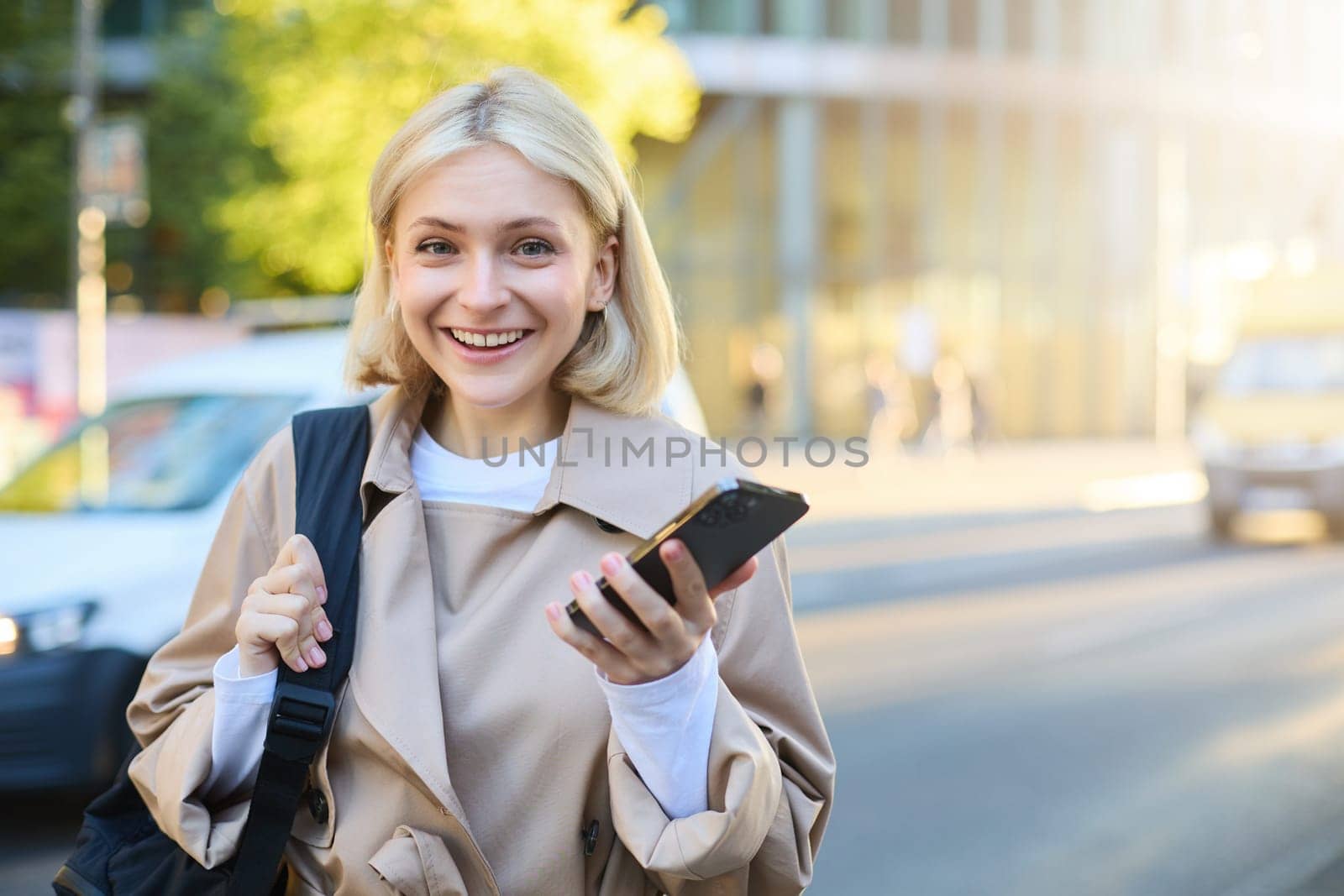 Close up portrait of young modern woman, standing on street, university student with mobile phone, looking happy and smiling at camera, using smartphone map app.