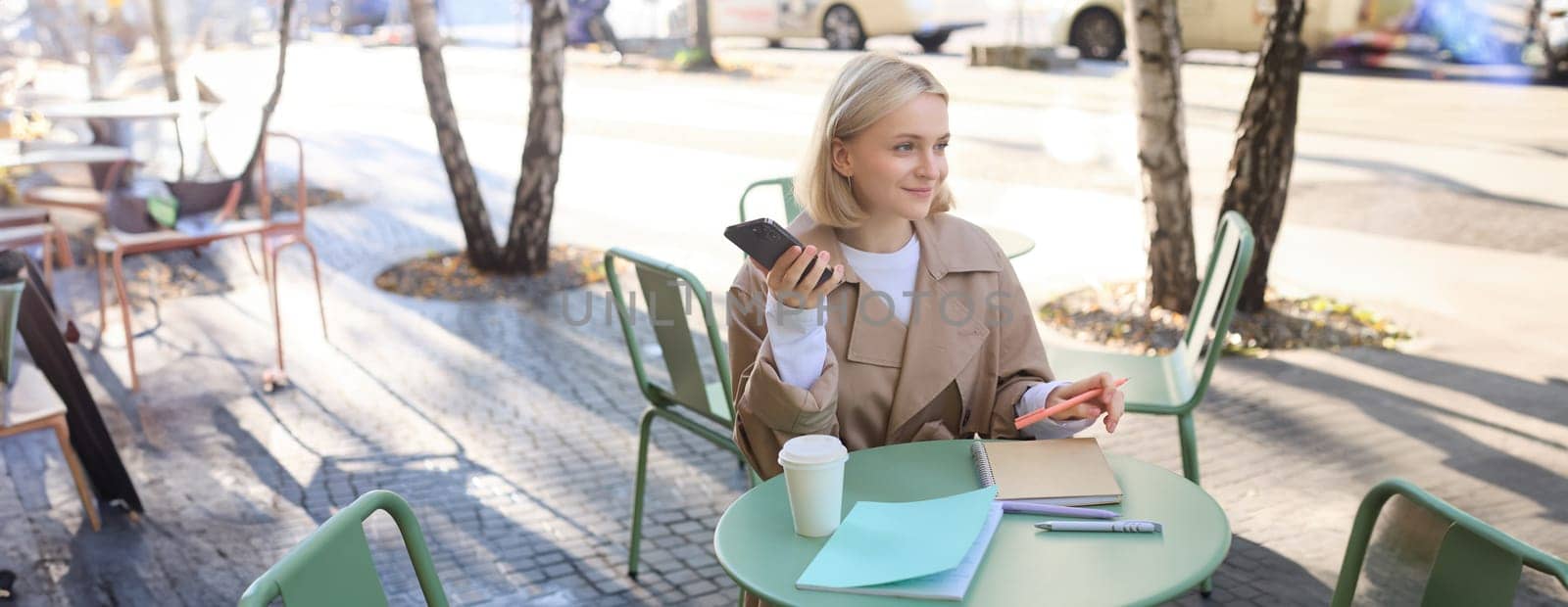Portrait of young woman studying, sitting in outdoor cafe with smartphone and journals, doing homework, wearing trench coat on sunny day by Benzoix