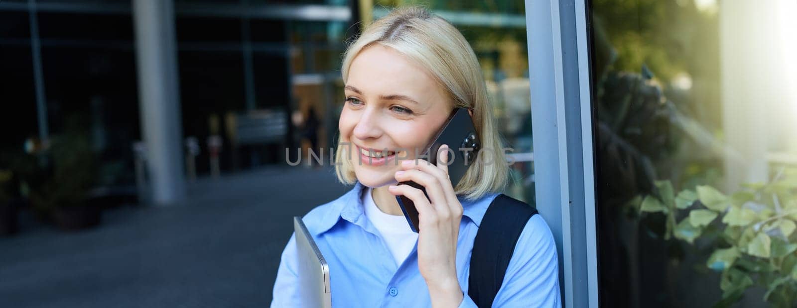 Portrait of young modern woman, office manager near building, standing outside with backpack, laptop, talking on mobile phone, chatting on smartphone. Lifestyle and communication concept