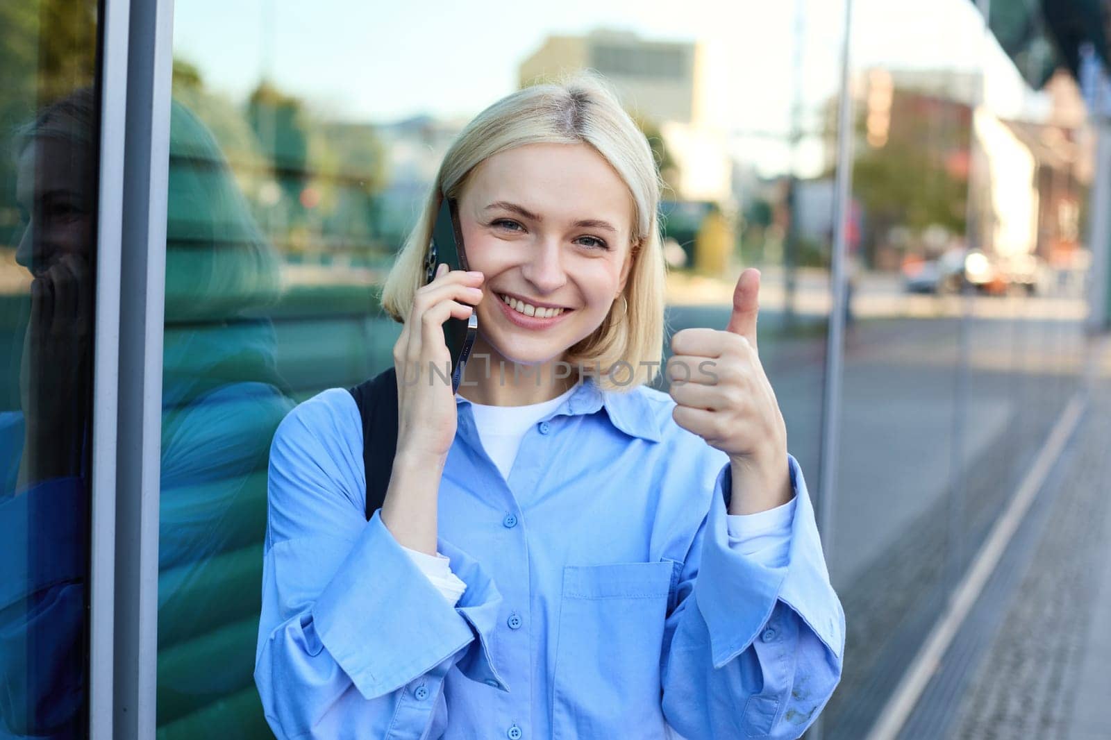 Close up portrait of blond smiling woman with backpack, standing on street near office building, showing thumb up in approval, recommending smth good by Benzoix